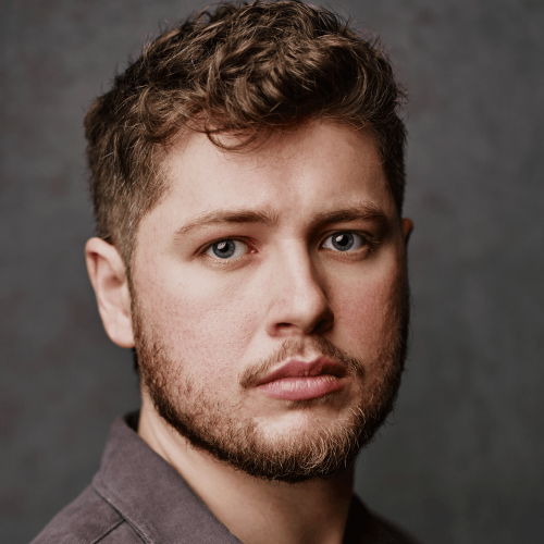 A white man with short brown hair with a curl on the top front of his head and a beard, looking directly at the camera. He is wearing a brown-gray top, standing in front of a dark gray wall.