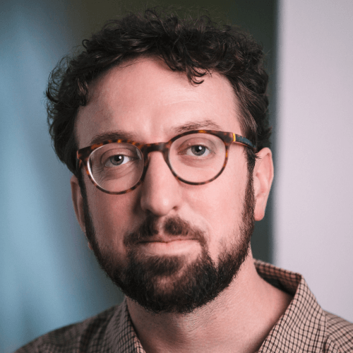 A white man with short curly brown hair and a beard, wearing glasses and a light and dark brown gingham shirt. He is in front of a white or gray wall.