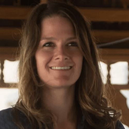 A white woman with long brown hair, wearing eyeliner and a dark top, smiling, looking away from the camera.