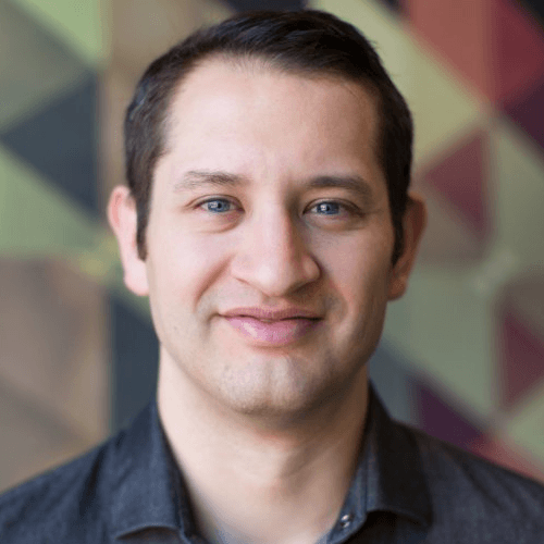 A man with short combed over brown hair, smiling in front of a multicolored and multi-triangle wall. He is wearing a denim top.