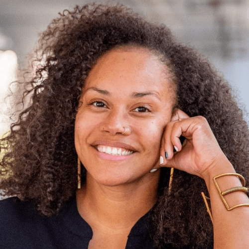 A Black woman with shoulder length curly black hair, smiling with her hand resting on the side of her face. She is wearing gold jewelry and a black top.