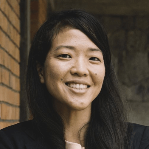 A grayscale or sepia photo. An Asian woman with long straight black hair, smiling in front of a brick wall. She is wearing a white shirt and a black jacket of sorts.