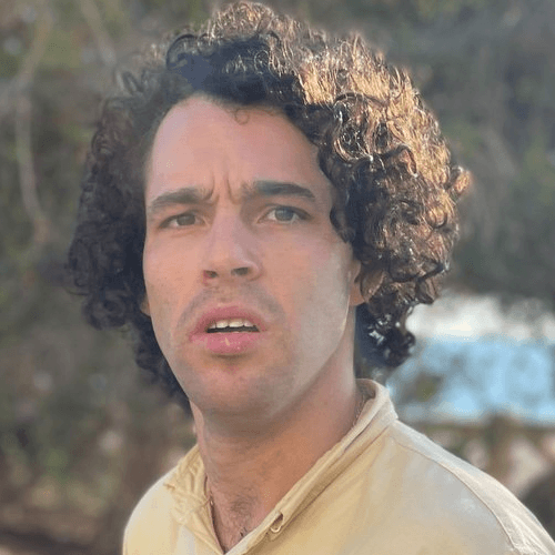 A man with chin length curly black hair, his mouth partially open as he looks at the camera. He is wearing a light colored shirt, and standing outside.