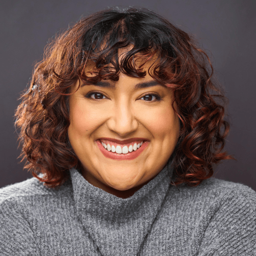 A Latinx person with chin length black curly hair, wearing a long sleeve pink shirt. They are holding a clear cane while sitting in front of a light orange wall