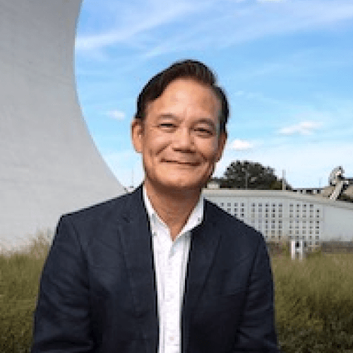 A Japanese man with short hair in a black jacket with a white shirt stands in front of a curved wall with blue sky behind him.