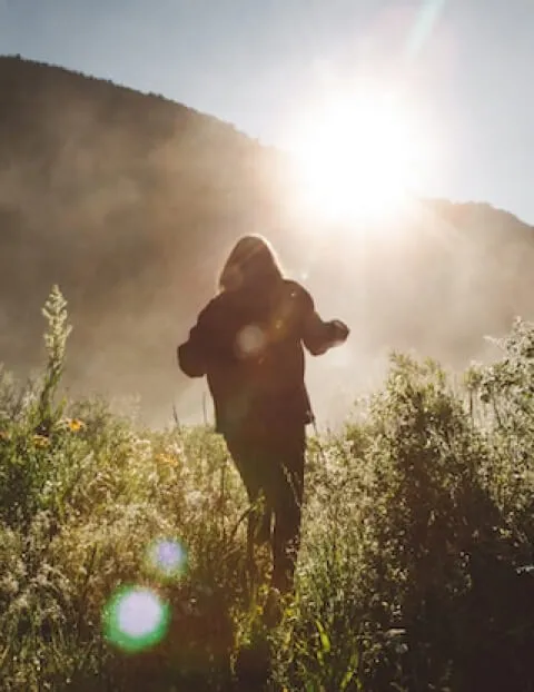 A hiker in a field of flowers with the sun peeking over a distant hill.