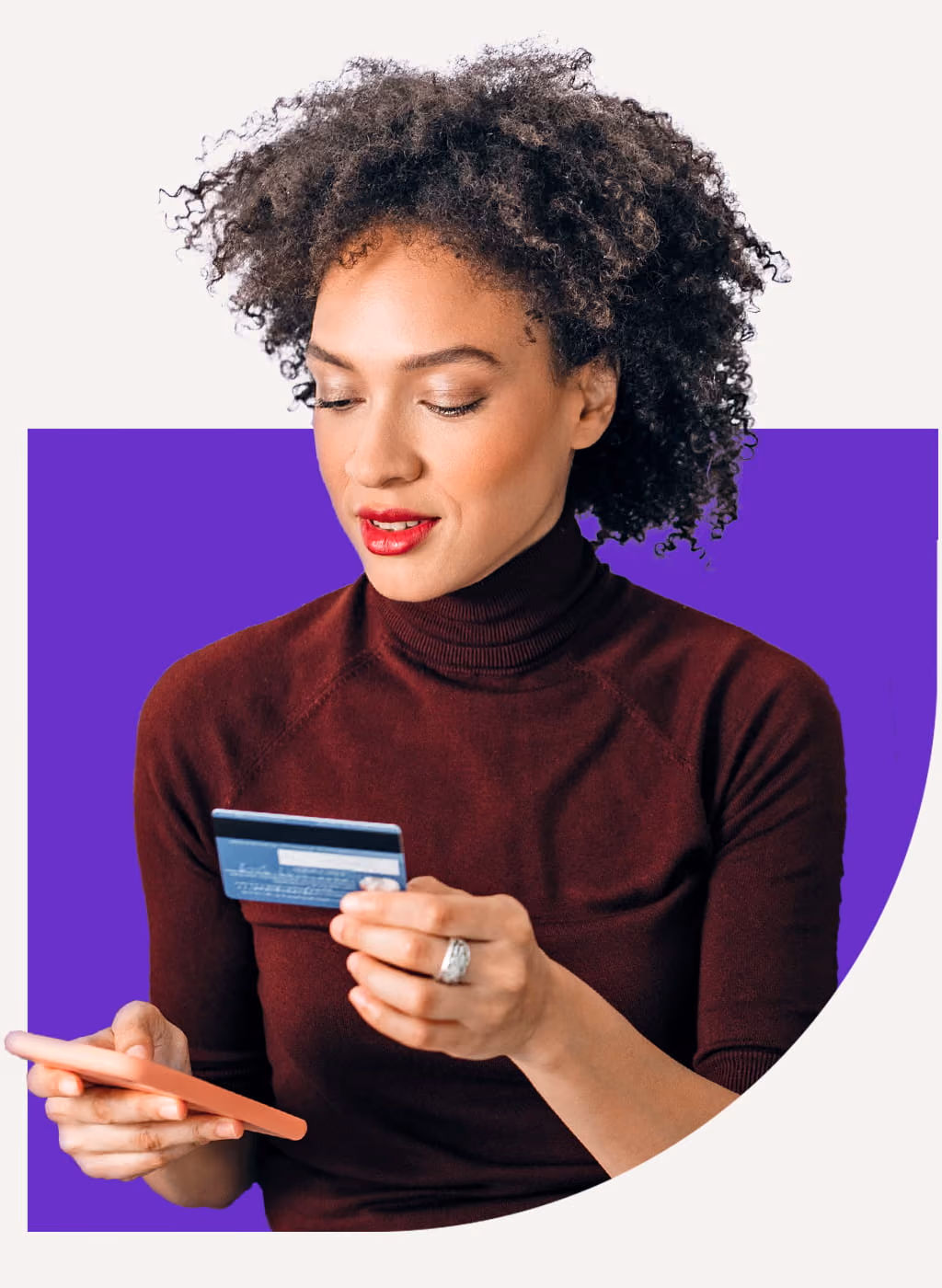 Woman with curly hair looking at a credit card while holding a smartphone.