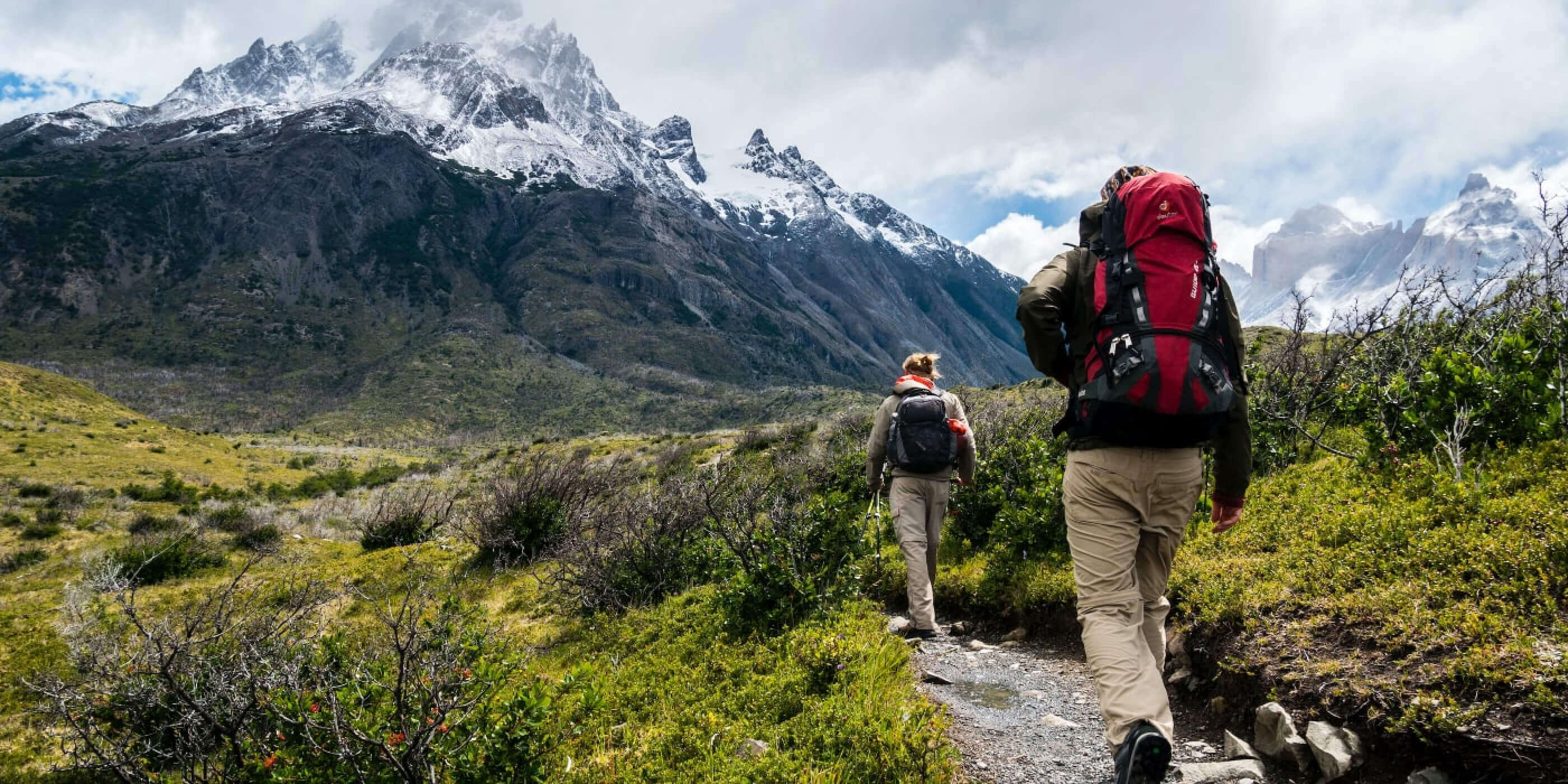 Two hikers walking a trail towards a snow-capped mountain.