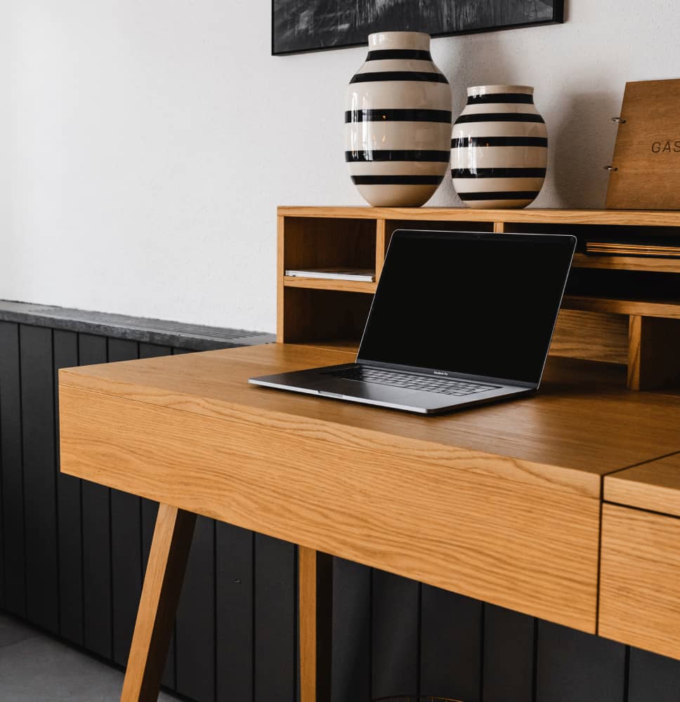 Wooden desk with open laptop and two black-and-white striped vases on a shelf against a white and black paneled wall.