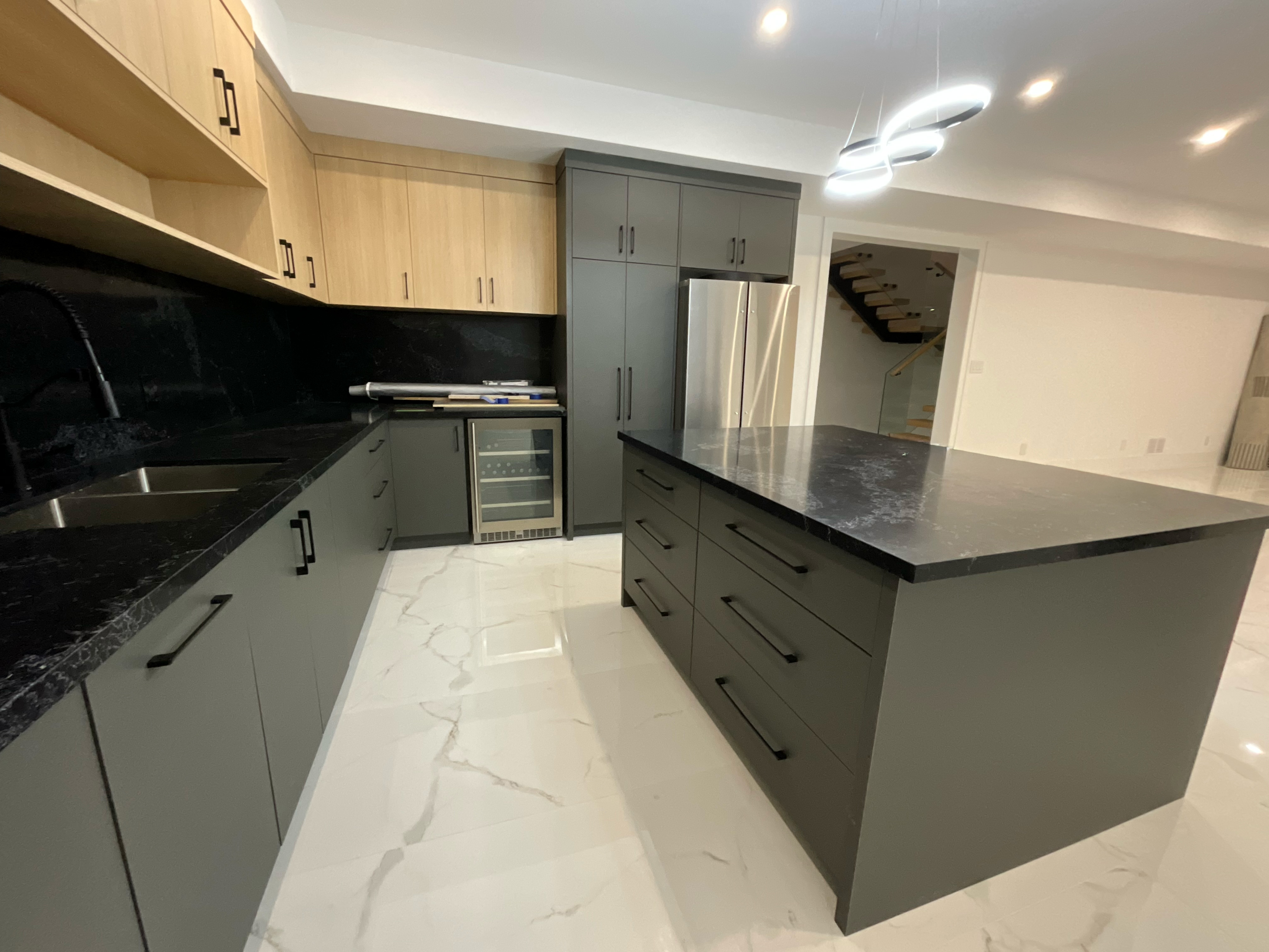 Modern kitchen with gray lower cabinets, light wood upper cabinets, black marble countertops, a central island, stainless steel refrigerator, and white marble floor tiles.
