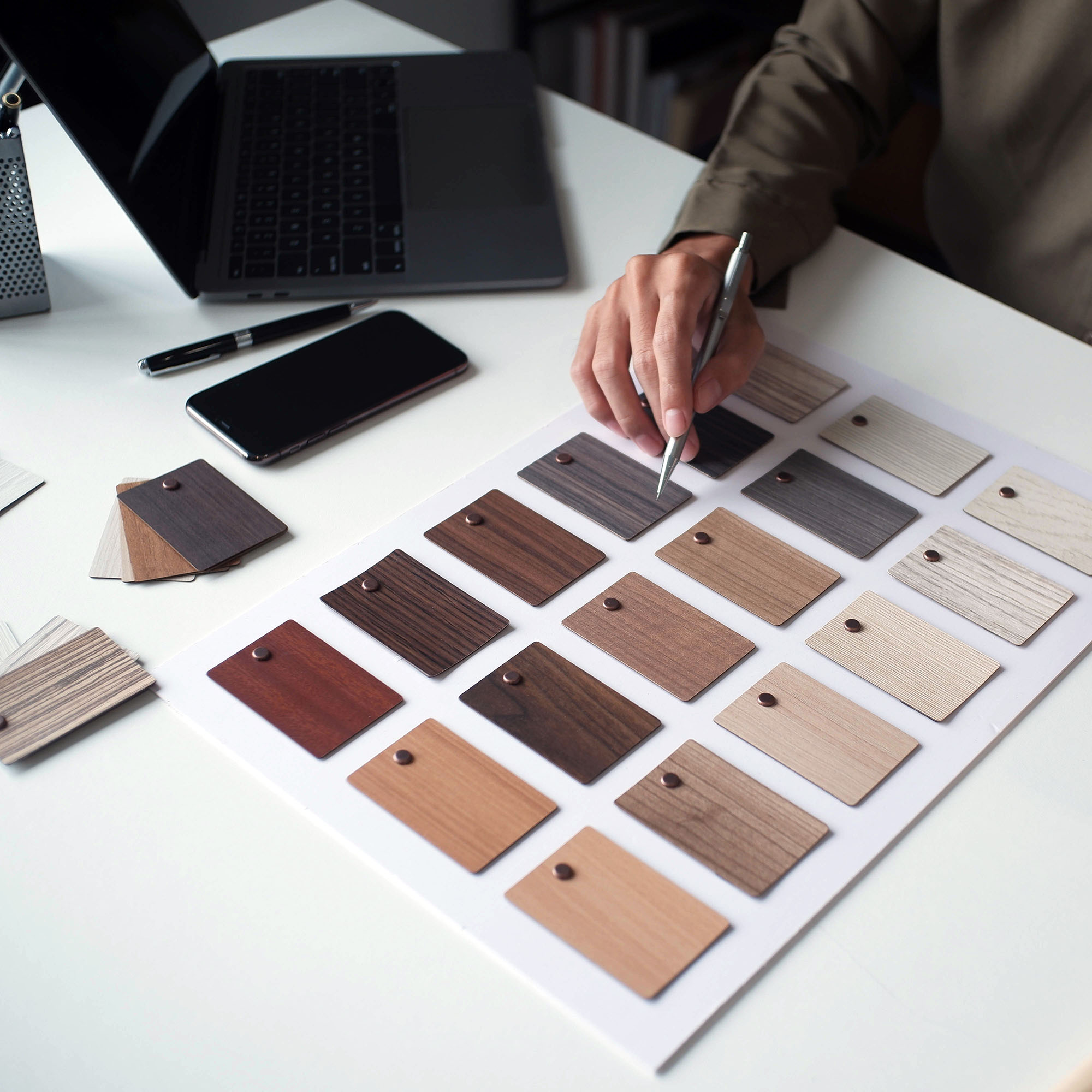 Person selecting a wood veneer sample from multiple wood finish swatches laid out on a white table next to a laptop and smartphone.