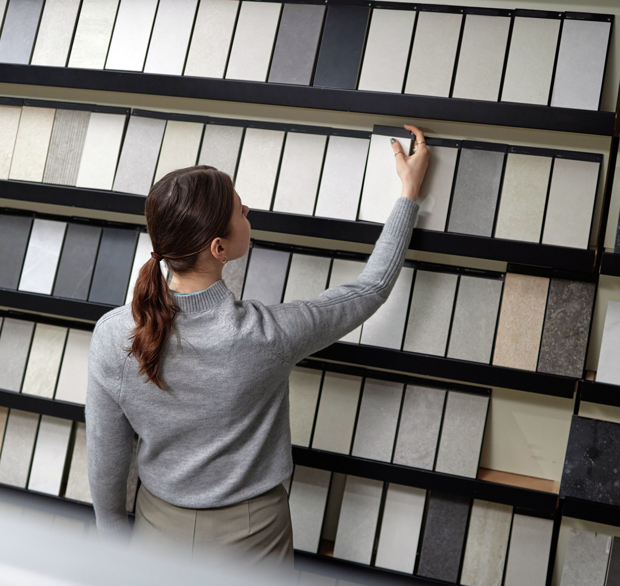 Woman in gray sweater selecting a tile sample from a display wall of various stone and ceramic tiles.