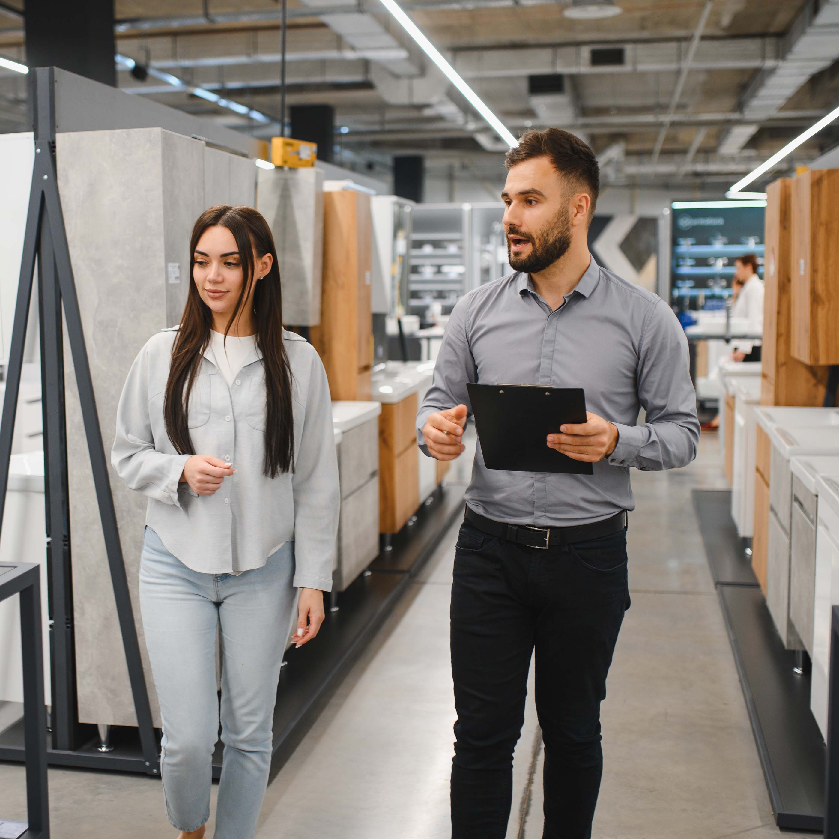 A man holding a clipboard talks to a woman as they walk through a modern showroom with kitchen cabinets and countertops.