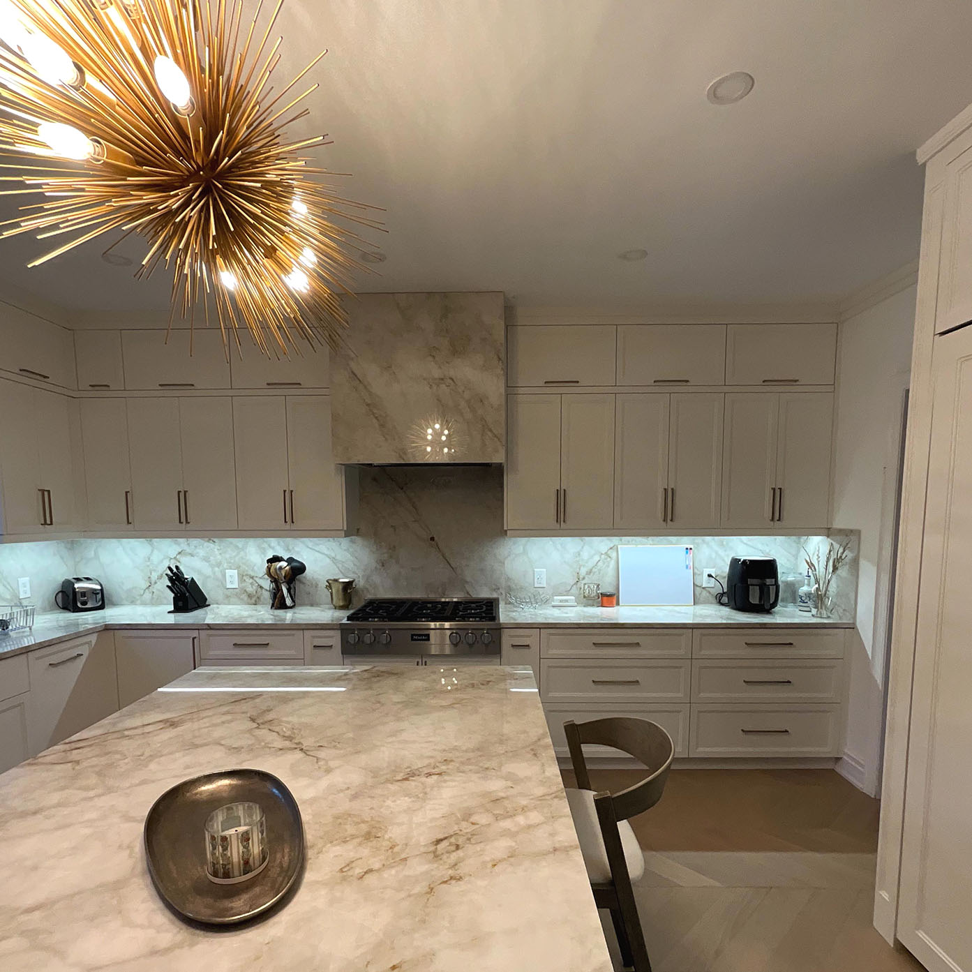 Modern kitchen with white cabinetry, marble countertops, a large island, and a distinctive gold starburst chandelier.
