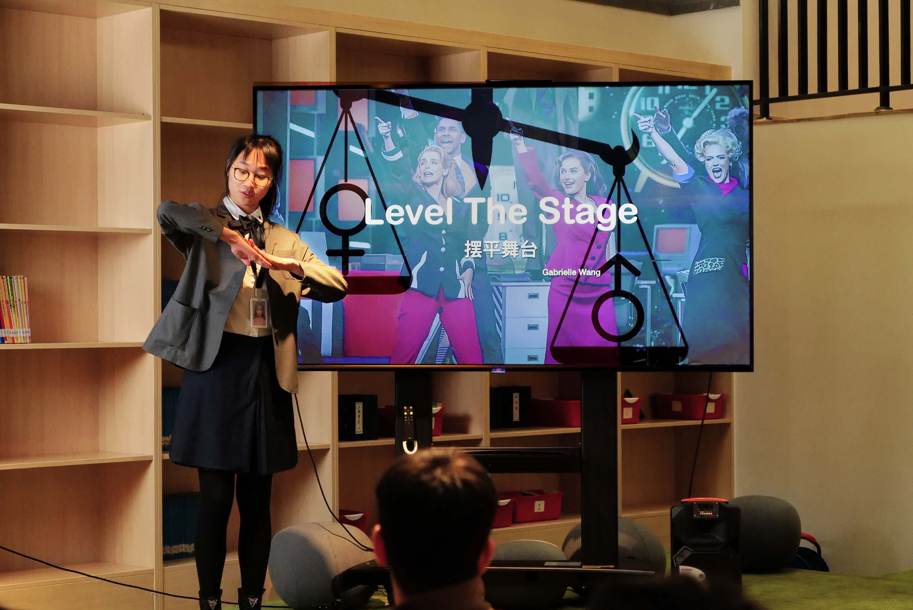 A student in uniform is giving a presentation in front of a large digital screen. The screen displays the title "Level The Stage" along with the presenter's name and an image of people celebrating. The student is gesturing with her hands as she explains, and the setting appears to be a classroom or lecture space.