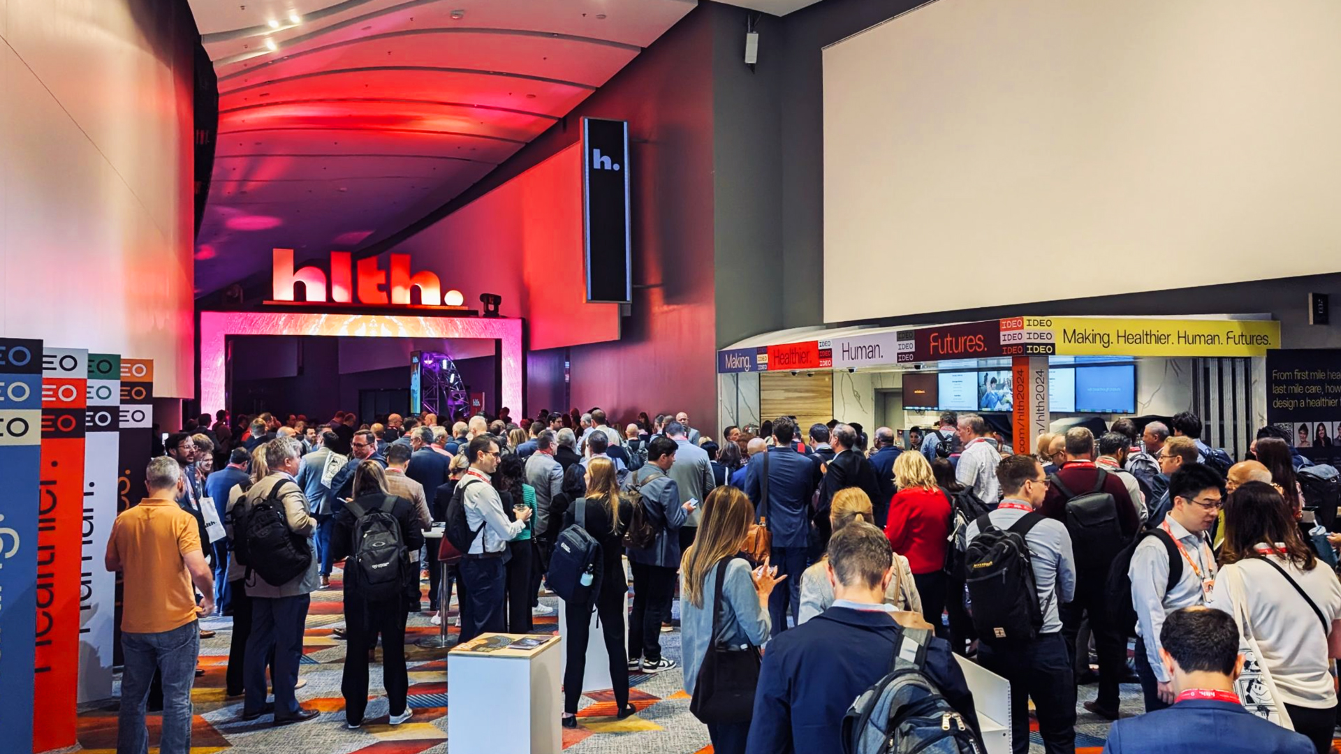 A large crowd of attendees gathers in a conference hallway illuminated by vibrant red and pink lighting. The "hlth." logo is prominently displayed above the entrance to a large event space. Booths and banners in the background display text such as "Making. Healthier. Human. Futures." Attendees are engaged in conversations and networking, with some holding bags and wearing name badges. The atmosphere is energetic and professional.