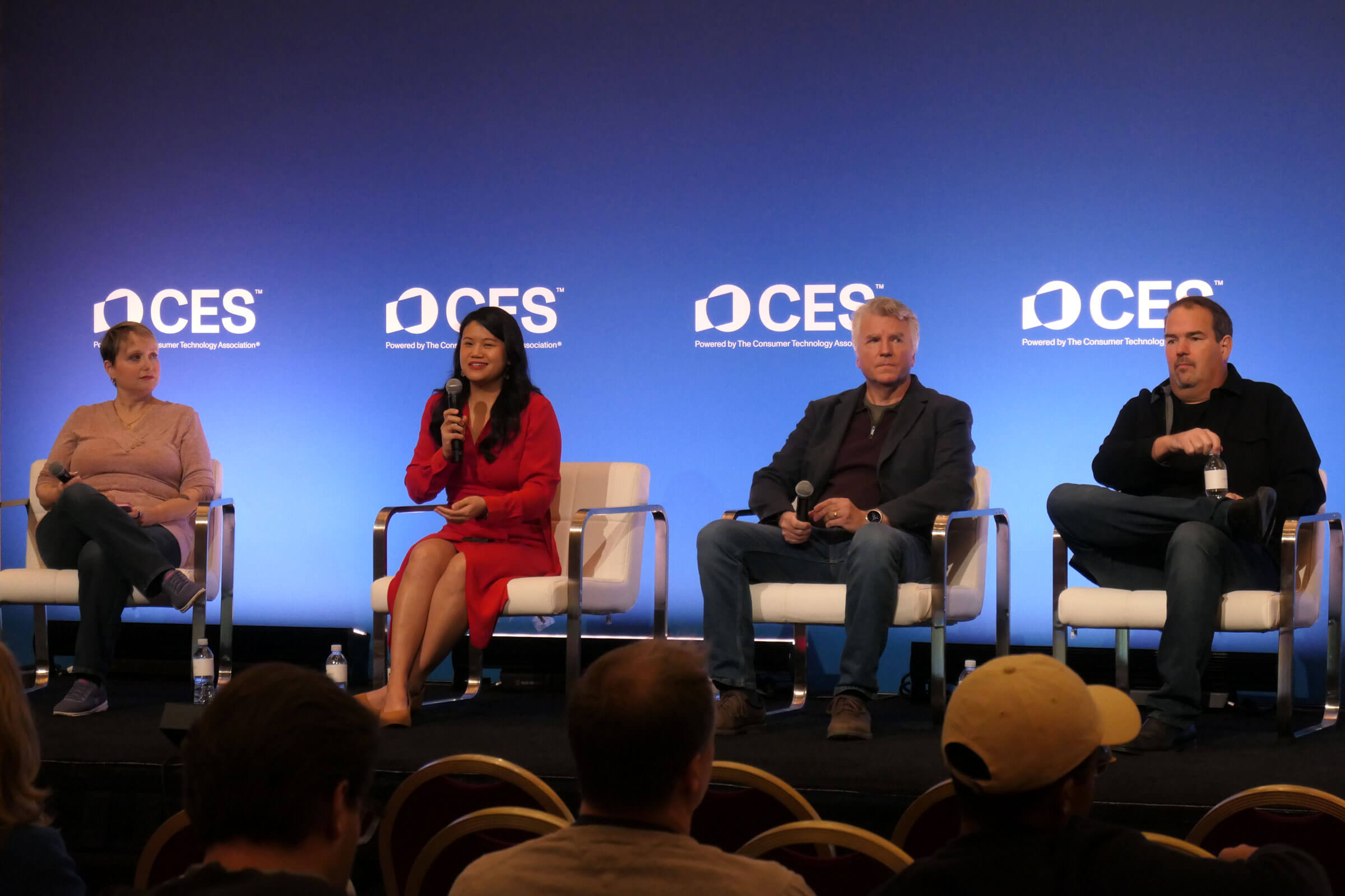CES panel discussion: A panel of four speakers on stage with a blue backdrop featuring the CES logo, engaged in a conversation while seated on white chairs. One speaker, dressed in a red dress, holds a microphone.