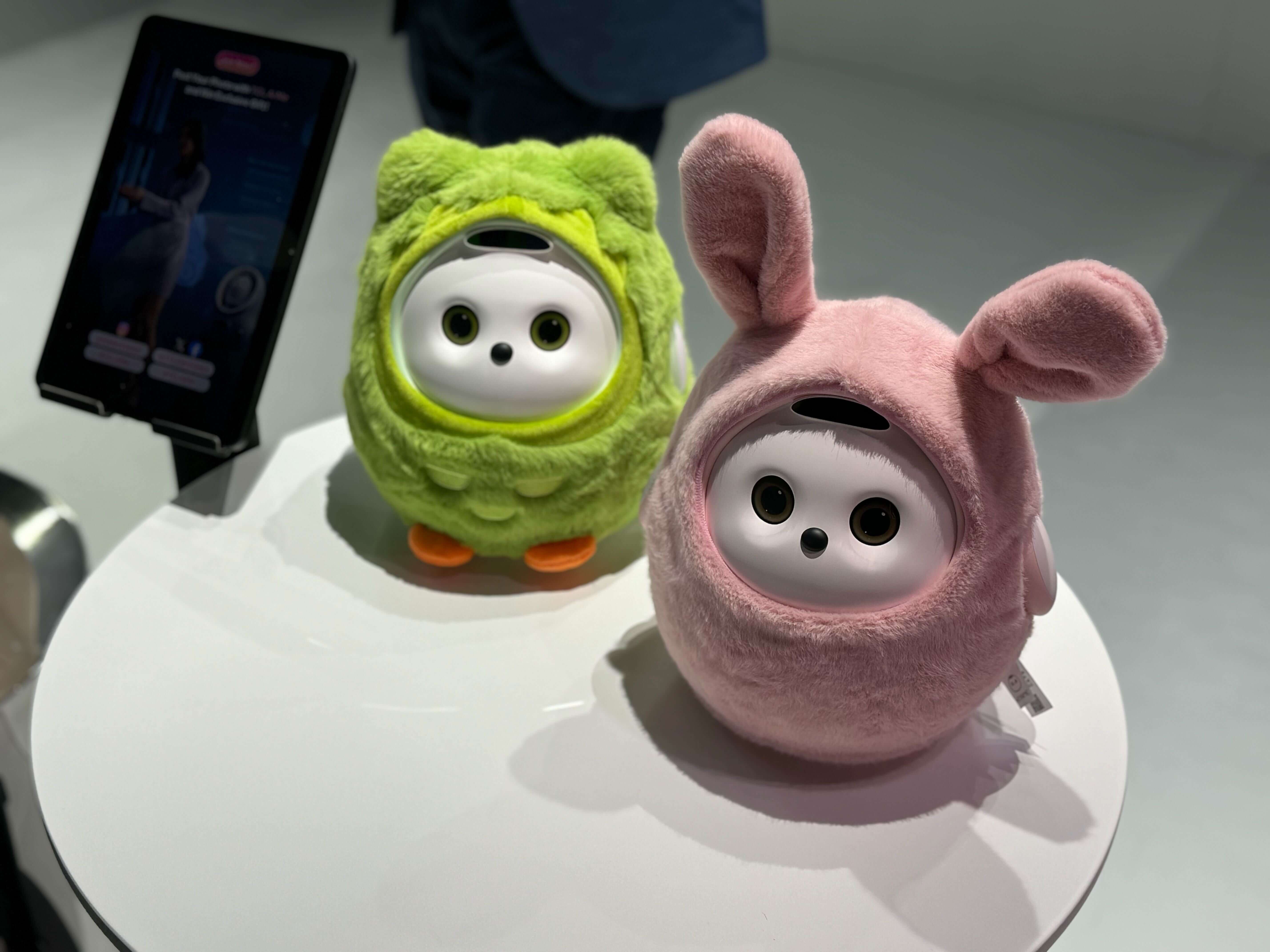 Cute robotic companions at CES: Two small, plush-covered robots on a white table. One is green with owl-like features, and the other is pink with bunny ears, each with expressive round eyes.