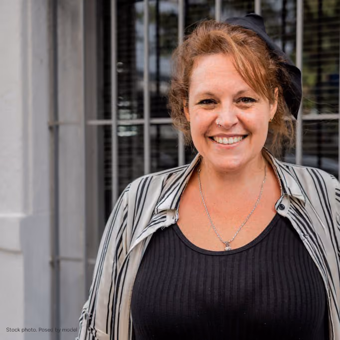 Smiling woman with reddish hair wearing a striped shirt over a black top standing outdoors in front of a window with bars.