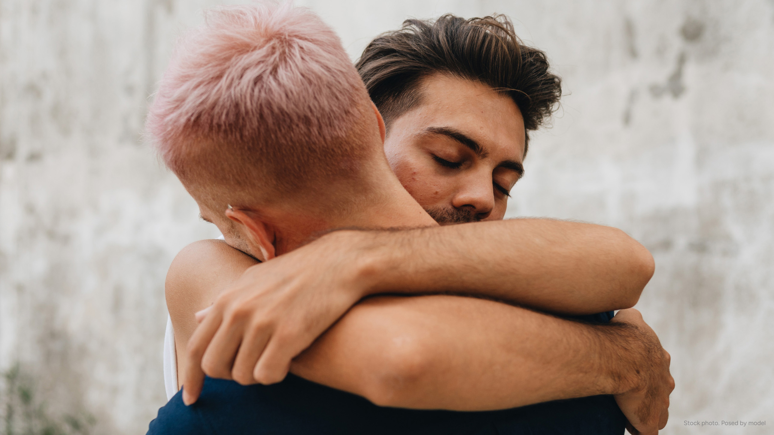 Two men hugging each other tightly, one with pink hair and the other with brown hair and a beard.