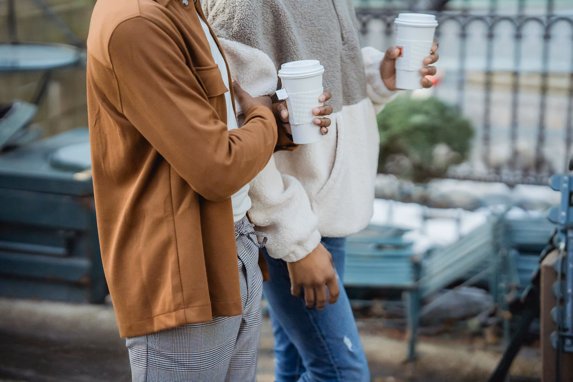Two people holding takeaway coffee cups outdoors, dressed in casual warm clothing.