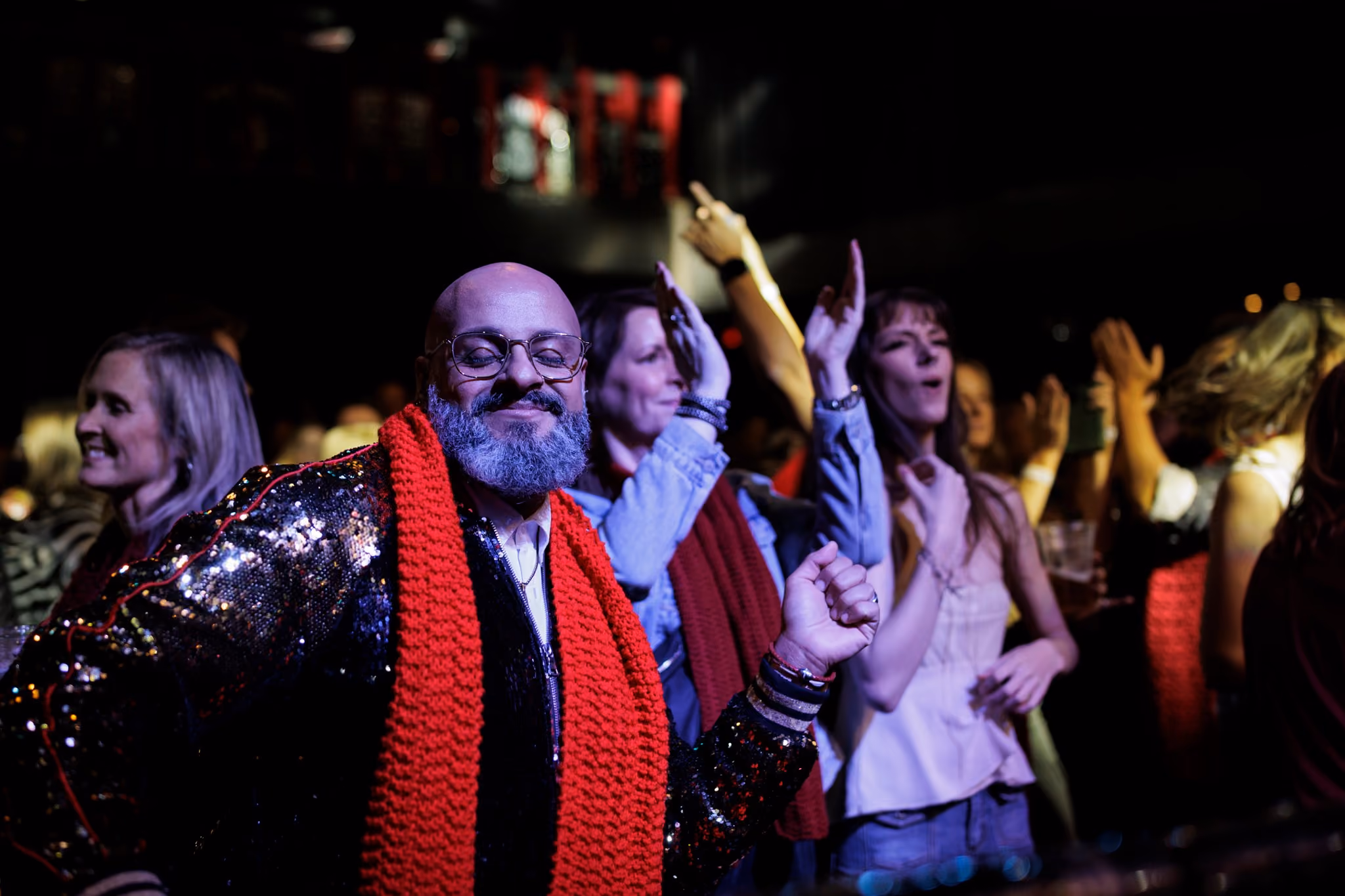 Man with glasses and gray beard wearing shiny jacket and red scarf dancing in a lively crowd at night.