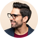 Smiling man with short dark hair and beard wearing black glasses and a dark blazer over a maroon shirt, facing left.