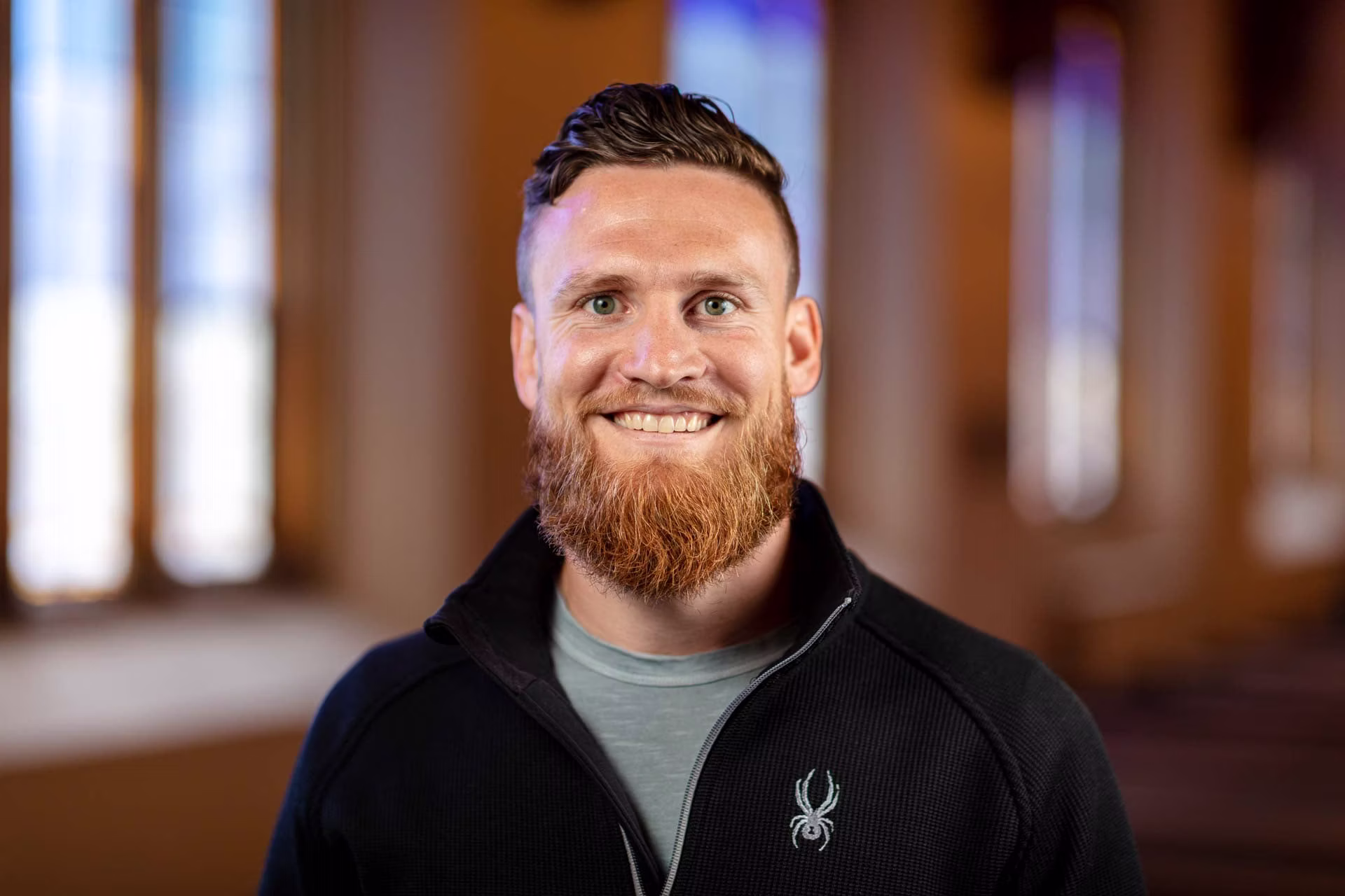 Smiling bearded man in black jacket and gray shirt indoors
