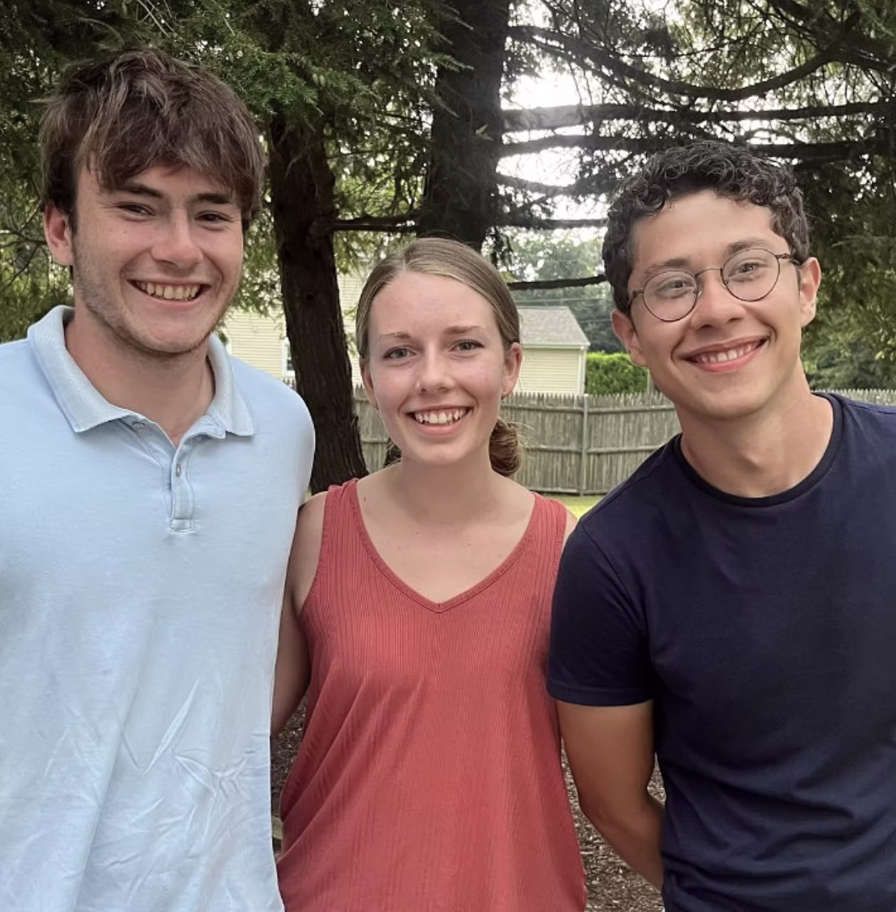 Three smiling friends standing together in a backyard with trees
