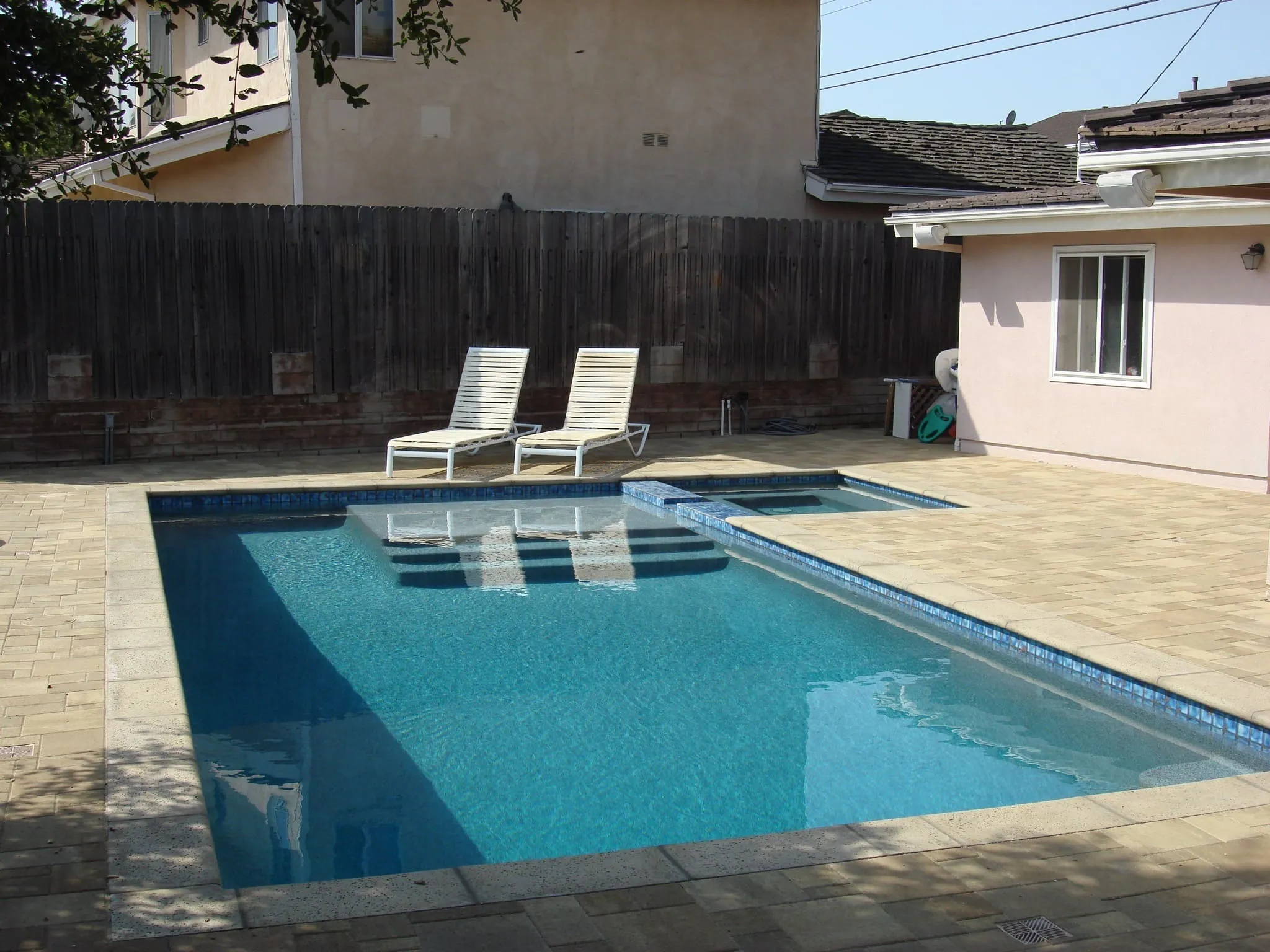 Rectangular backyard swimming pool with blue water and adjacent hot tub surrounded by beige stone tiles and two white lounge chairs.