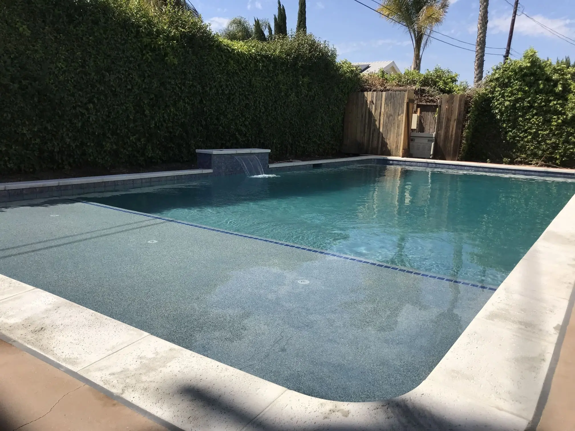 Rectangular swimming pool with clear blue water, a small waterfall feature, and surrounded by a white concrete deck and green hedges.