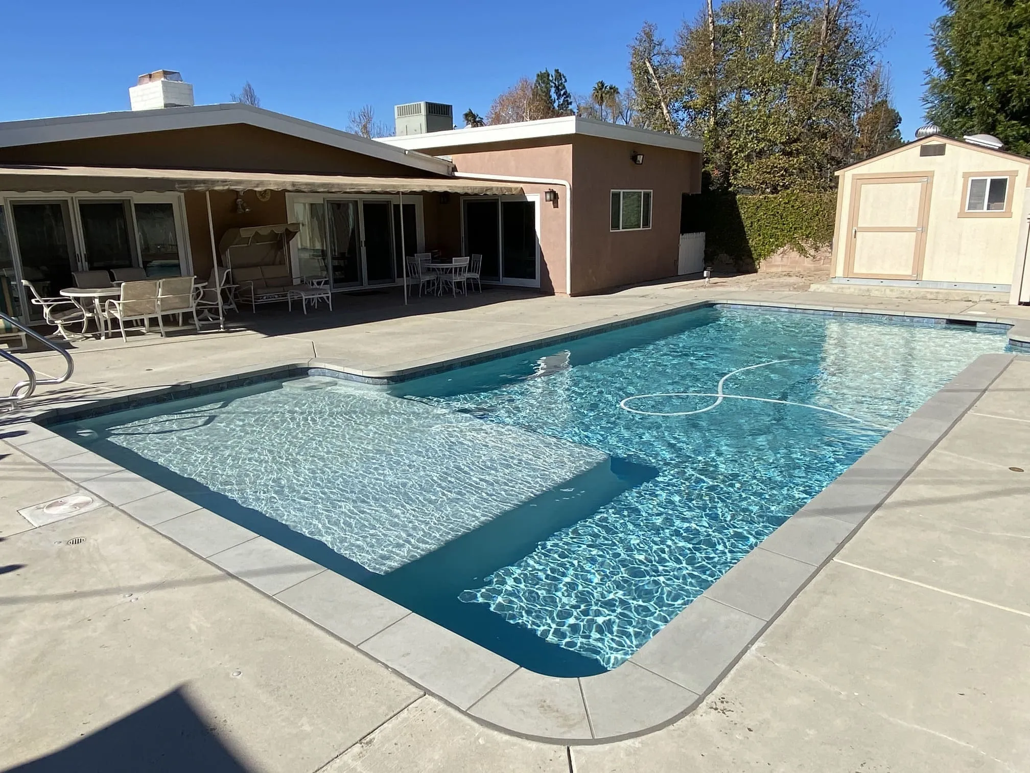 Outdoor swimming pool with clear blue water beside a beige house with patio furniture and a storage shed in the background.