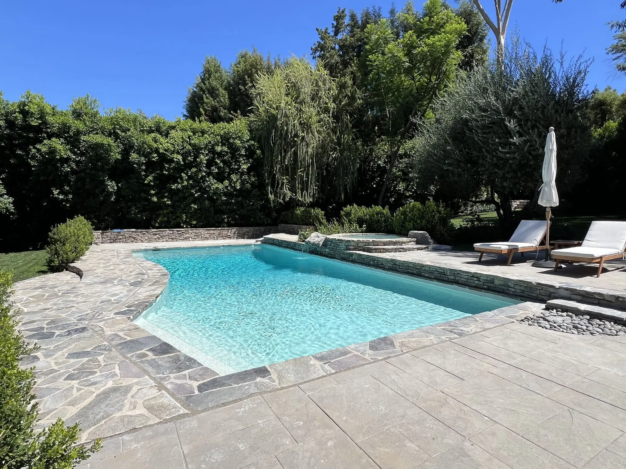 Outdoor swimming pool with clear blue water, surrounded by stone pavement, greenery, two white lounge chairs and a closed umbrella.