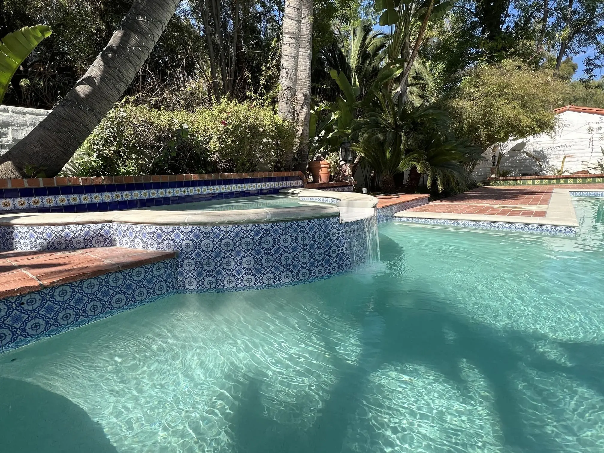 Outdoor swimming pool with a tiled edge, a small waterfall from a raised spa, surrounded by palm trees and greenery.