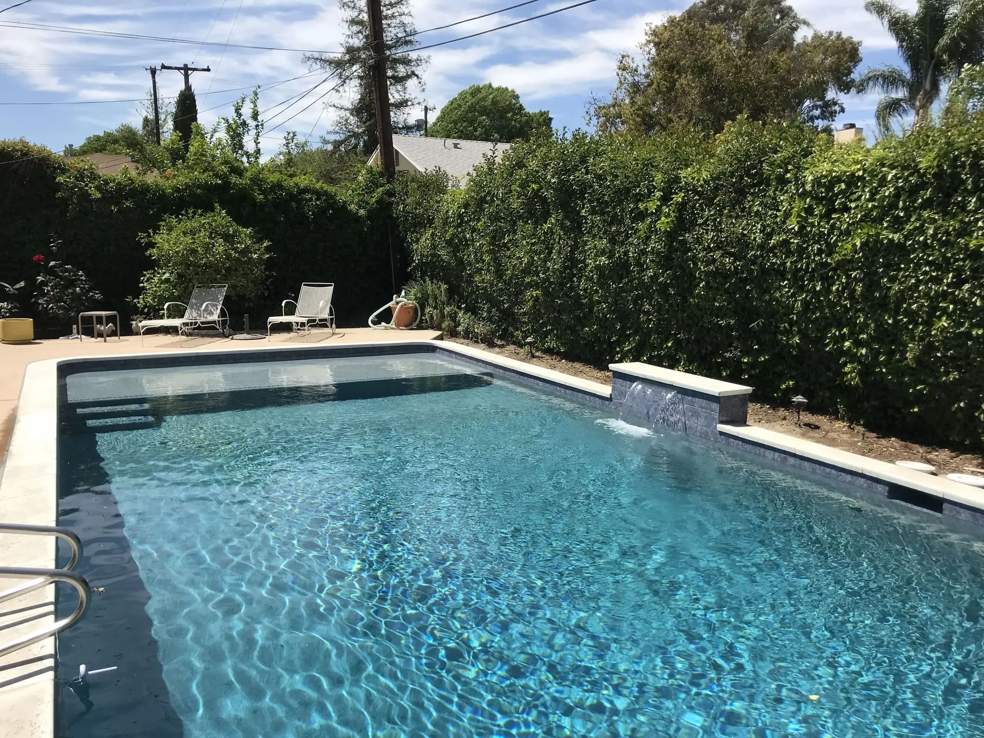 Clear blue swimming pool with water feature, surrounded by a hedge and lounge chairs on the pool deck.