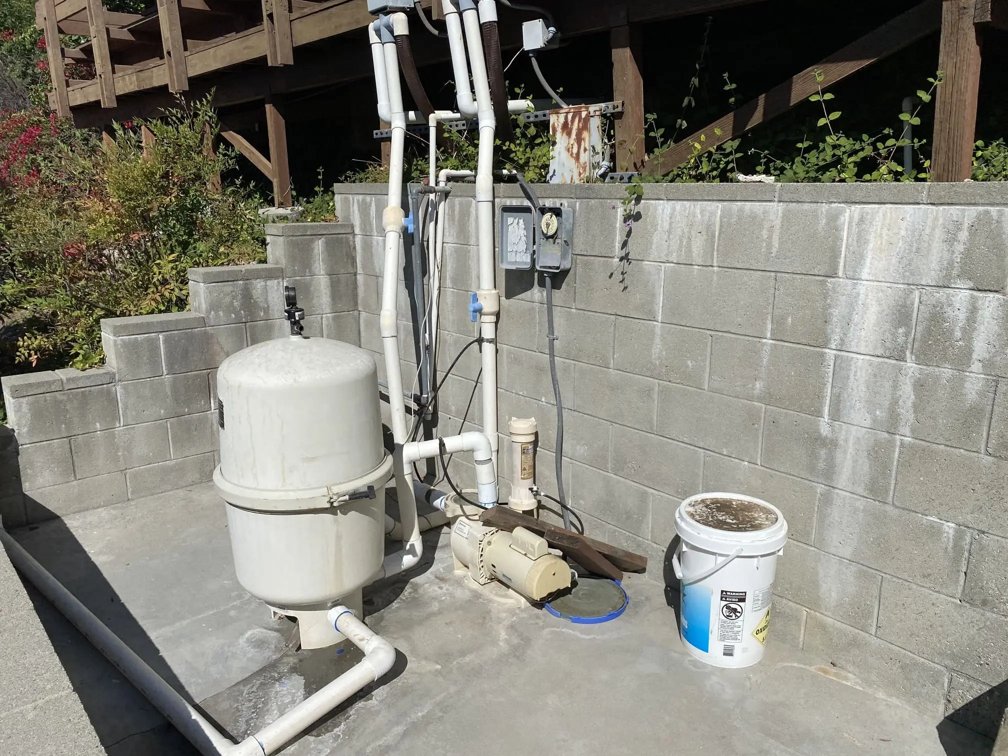 Outdoor pool equipment setup with a large white filter tank, pump, PVC pipes, and a white bucket on a concrete slab against a cinder block wall.