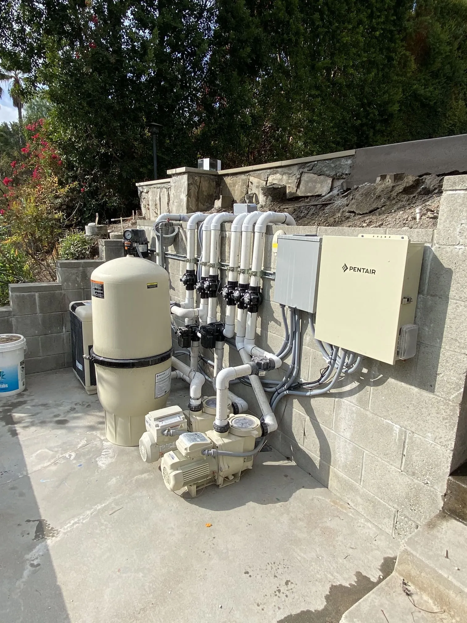 Outdoor pool pump system with white PVC pipes, two motors, a large filter tank, and a Pentair control box mounted on a cinder block wall.