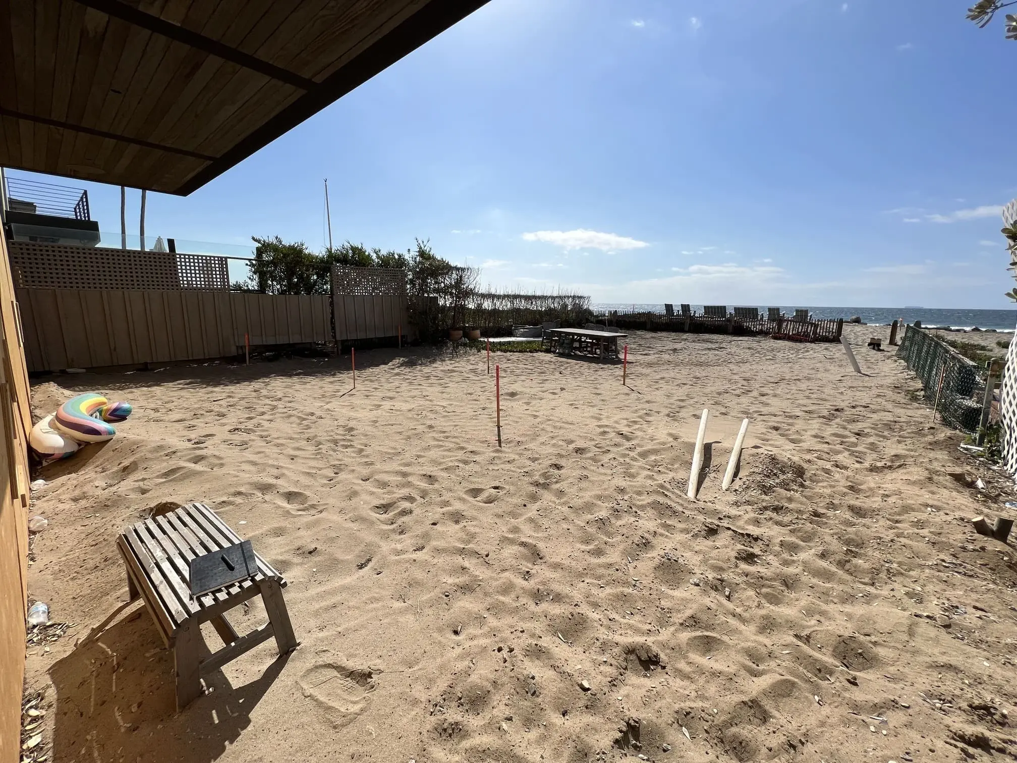 Sandy backyard area with a wooden bench, pipes sticking out of sand, and a view of the ocean under a clear sky.