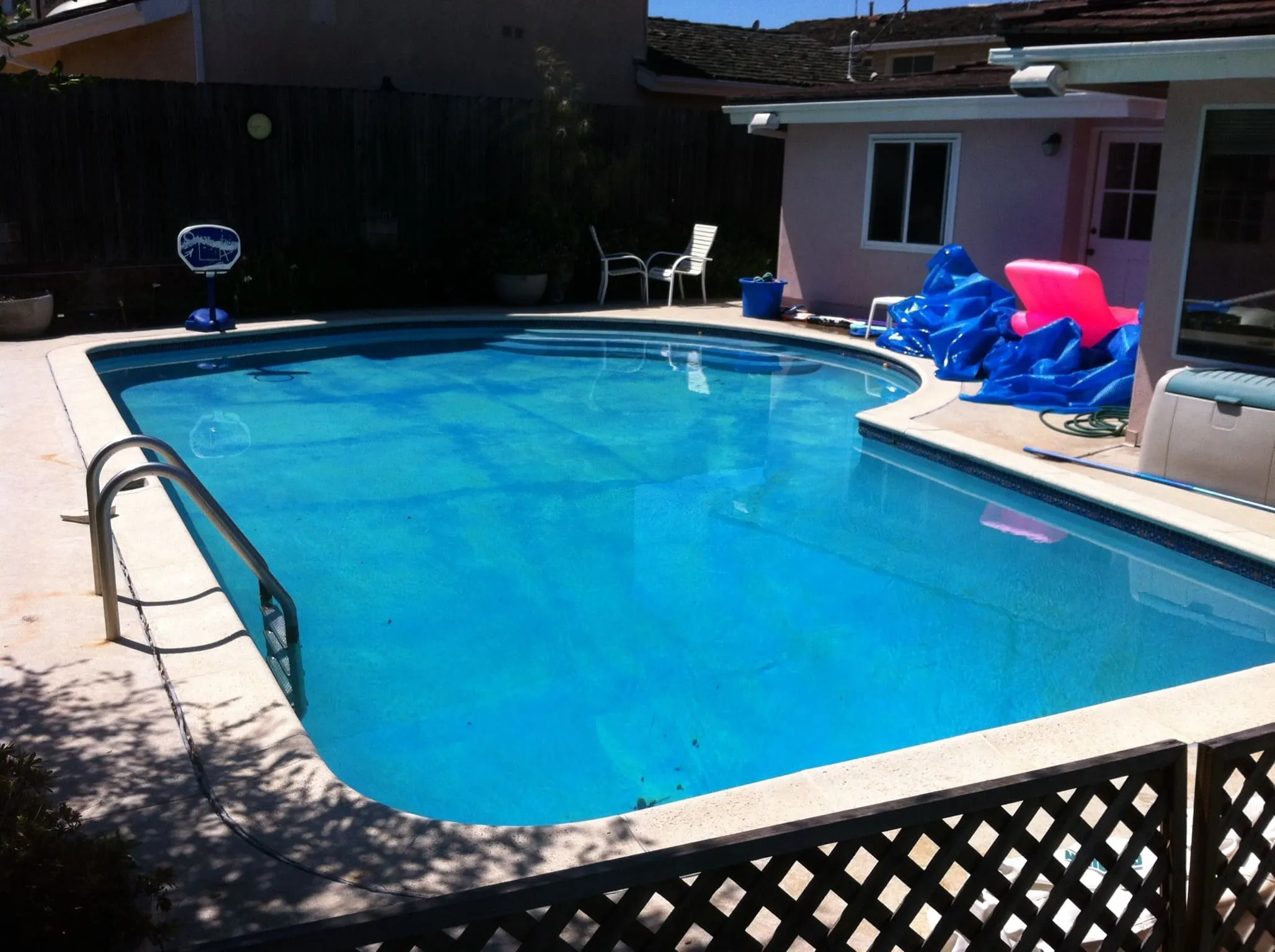 Backyard swimming pool with clear blue water, metal ladder, white patio chairs, and pool toys near a house.