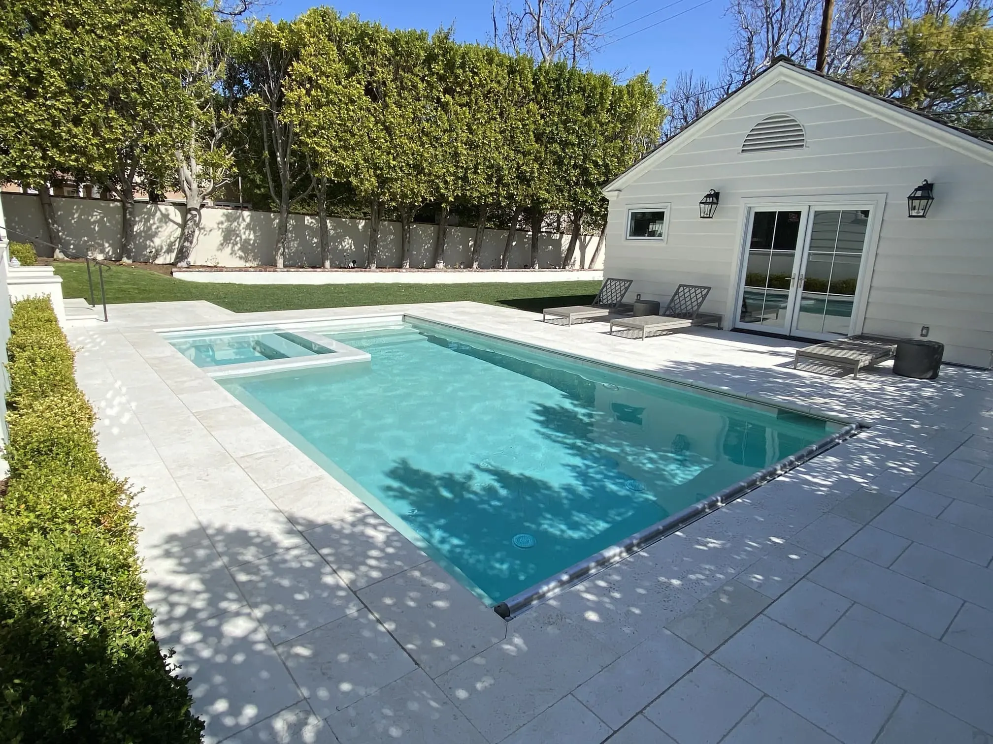 Modern backyard with a rectangular swimming pool, attached square hot tub, white tiled deck, lounge chairs, and tall green hedges along the fence.