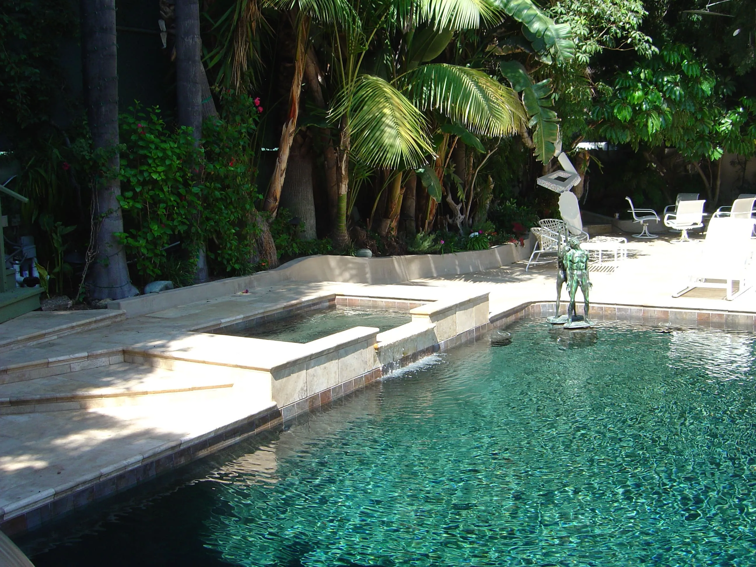 Swimming pool with clear turquoise water, a small adjacent hot tub, surrounded by tropical plants and white patio furniture, and a sculpture near the pool edge.