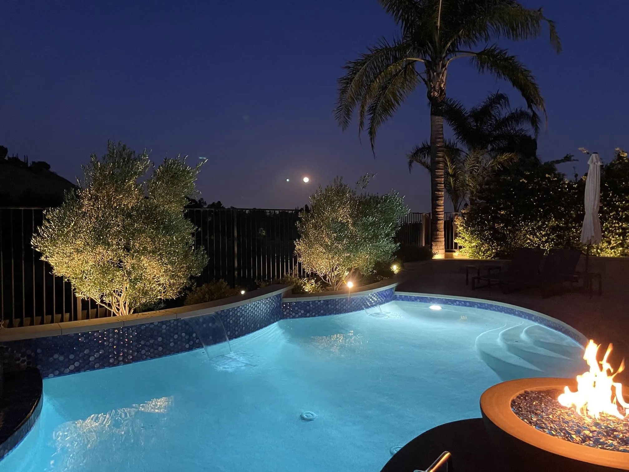 Illuminated backyard swimming pool at night with fire pit, palm trees, and moon in the sky.