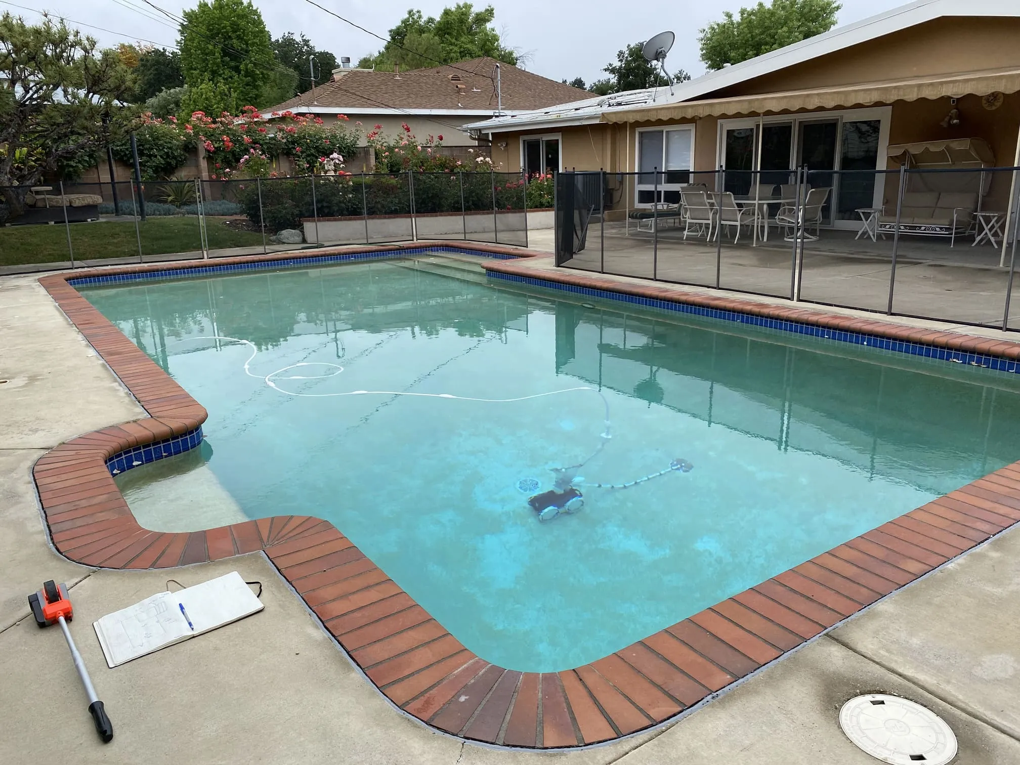 Residential swimming pool with a robotic pool cleaner in the water and patio furniture behind a black safety fence.