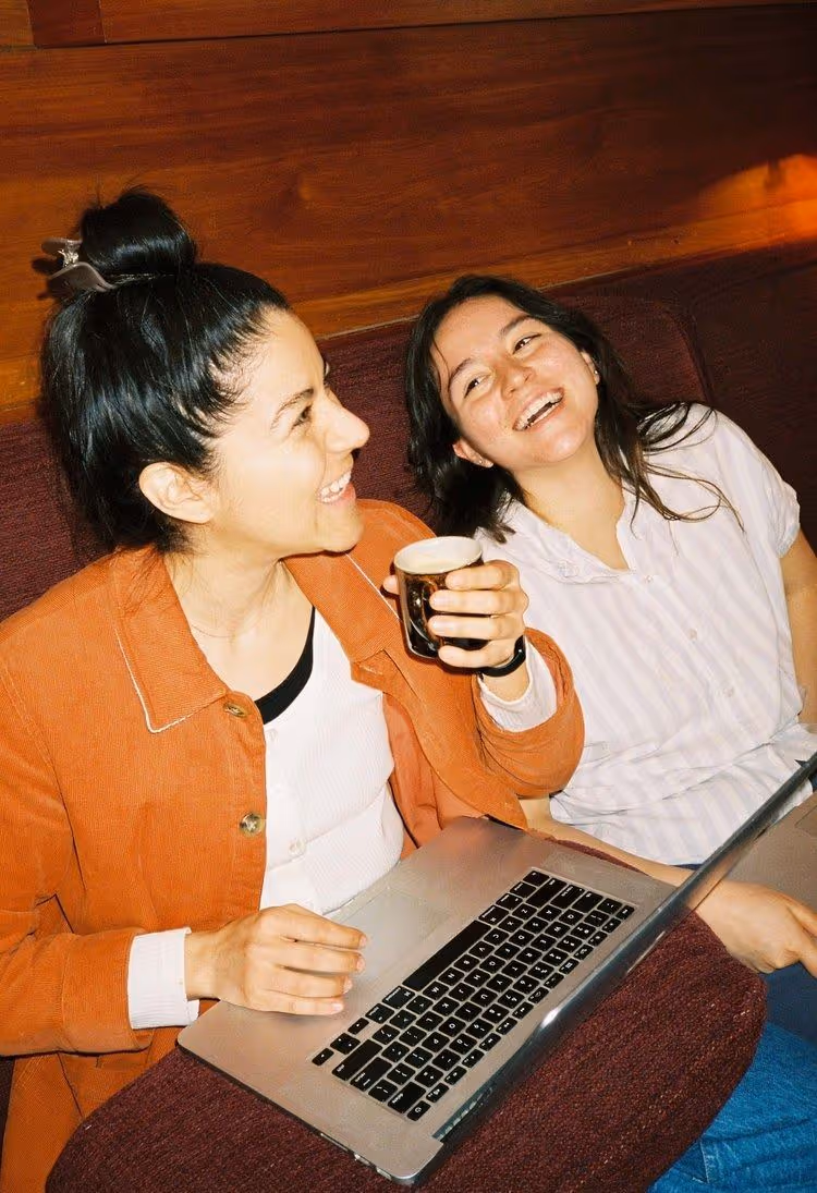 Two girls laughing and holding a cup and laptop. Brown cafe.