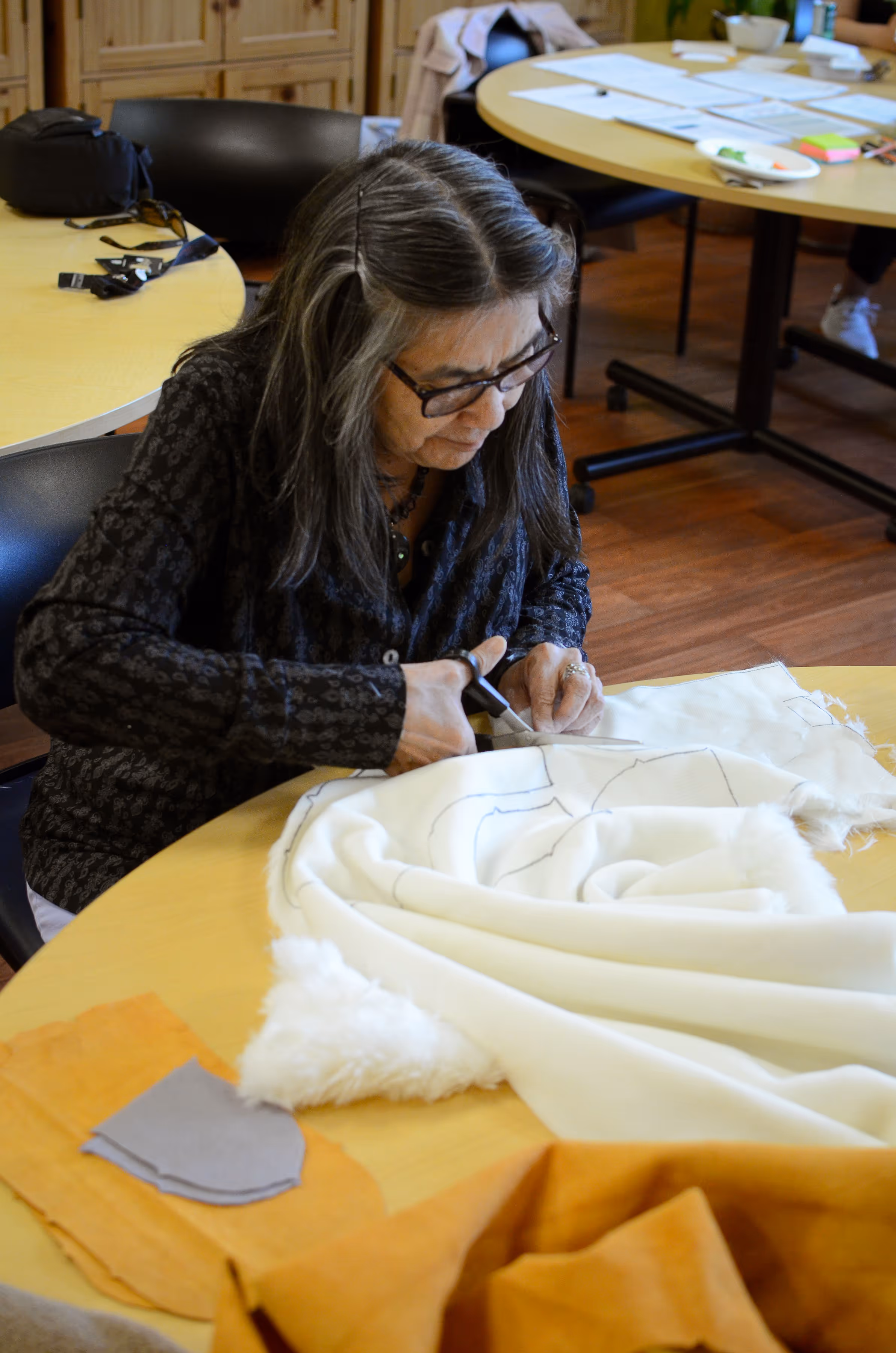 Elderly woman with glasses cutting white fabric with scissors at a table covered with sewing materials.