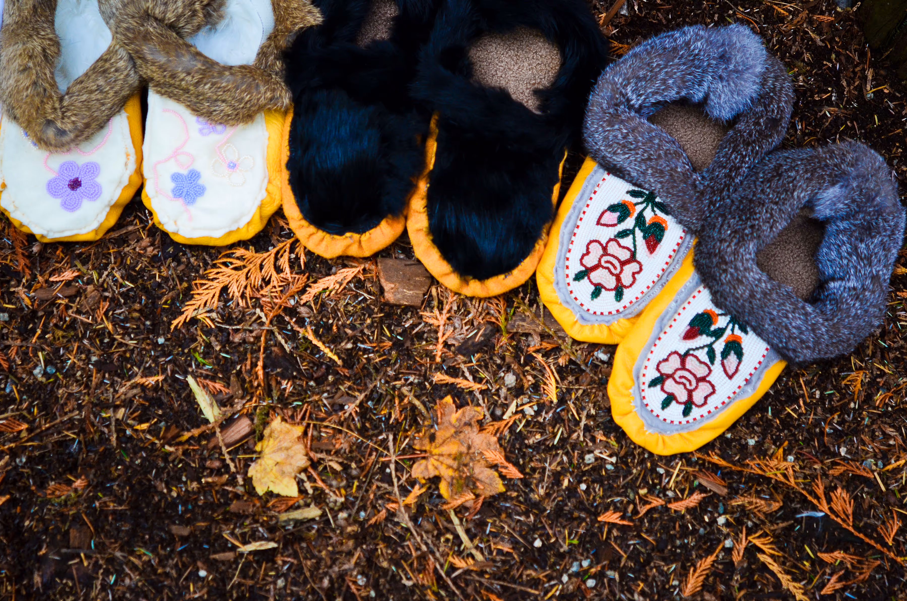 Three pairs of moccasins with different colored fur trims and embroidered floral designs laid on forest floor.