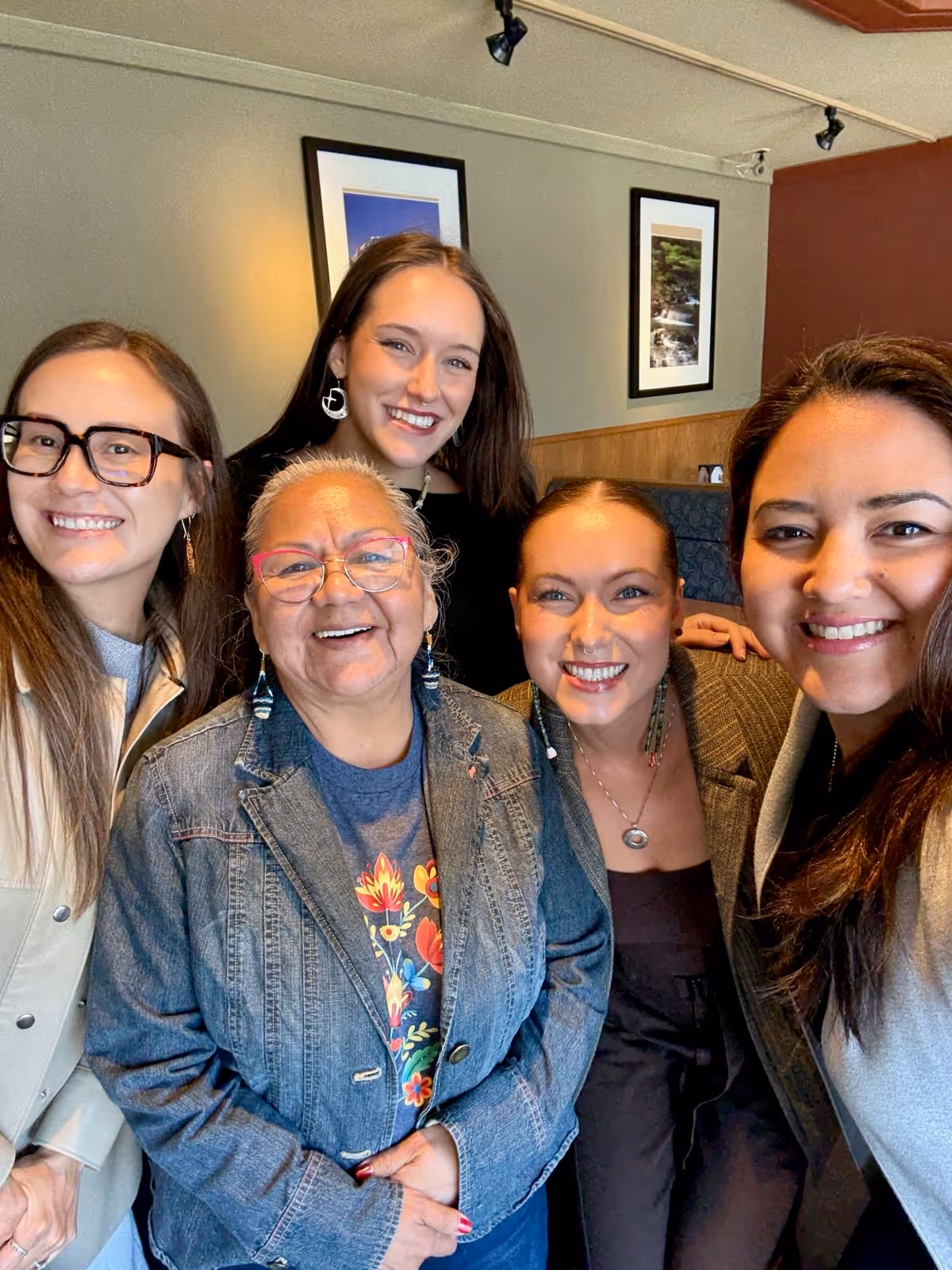 Five women smiling together in a cozy indoor setting with framed pictures on the wall behind them.