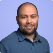 Smiling man with short hair and beard wearing a blue collared shirt against a light purple background.