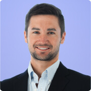 Smiling man with short dark hair wearing a dark blazer and light blue collared shirt against a light purple background.