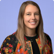 Smiling woman with long blonde hair wearing a colorful floral jacket over a black top against a blue background.
